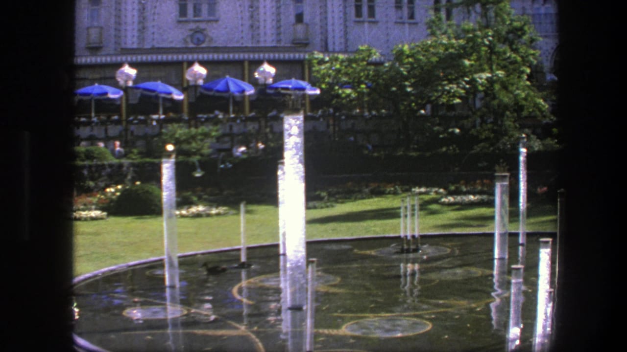 1975 Copenhagen home-movie frame with bright water jets in a circular pool, cafe-style blue umbrellas, greenery, and an ornate facade.