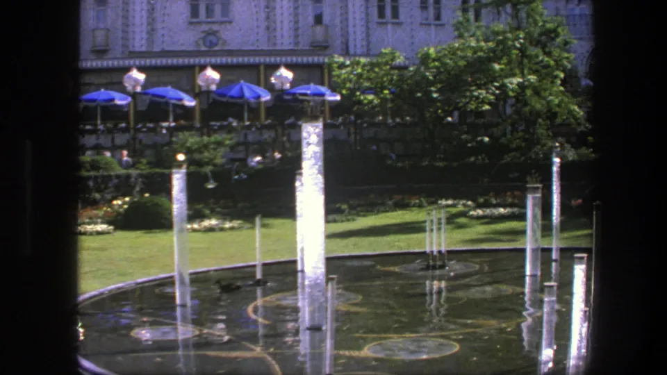 1975 Copenhagen home-movie frame with bright water jets in a circular pool, cafe-style blue umbrellas, greenery, and an ornate facade.