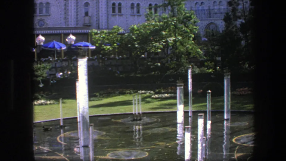 1975 Copenhagen 8mm frame looking through a dark edge at fountain columns, reflective water, lawns, trees, blue umbrellas, and distant people.