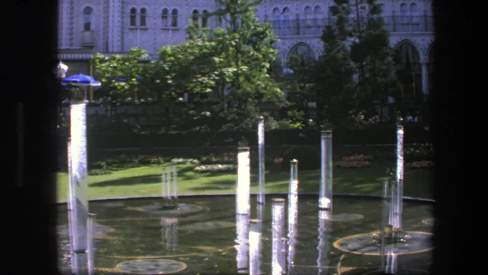1975 Copenhagen home-movie frame of several fountain jets, circular pool markings, greenery, blue umbrellas, and ornate architecture in bright daylight.