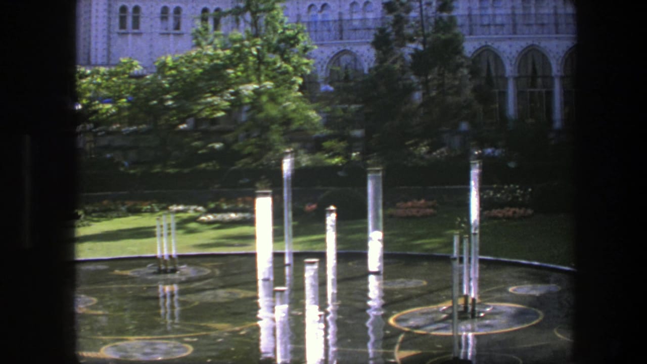 Reflecting pool with white columns, green lawn, trees, and pale arched building behind it.
