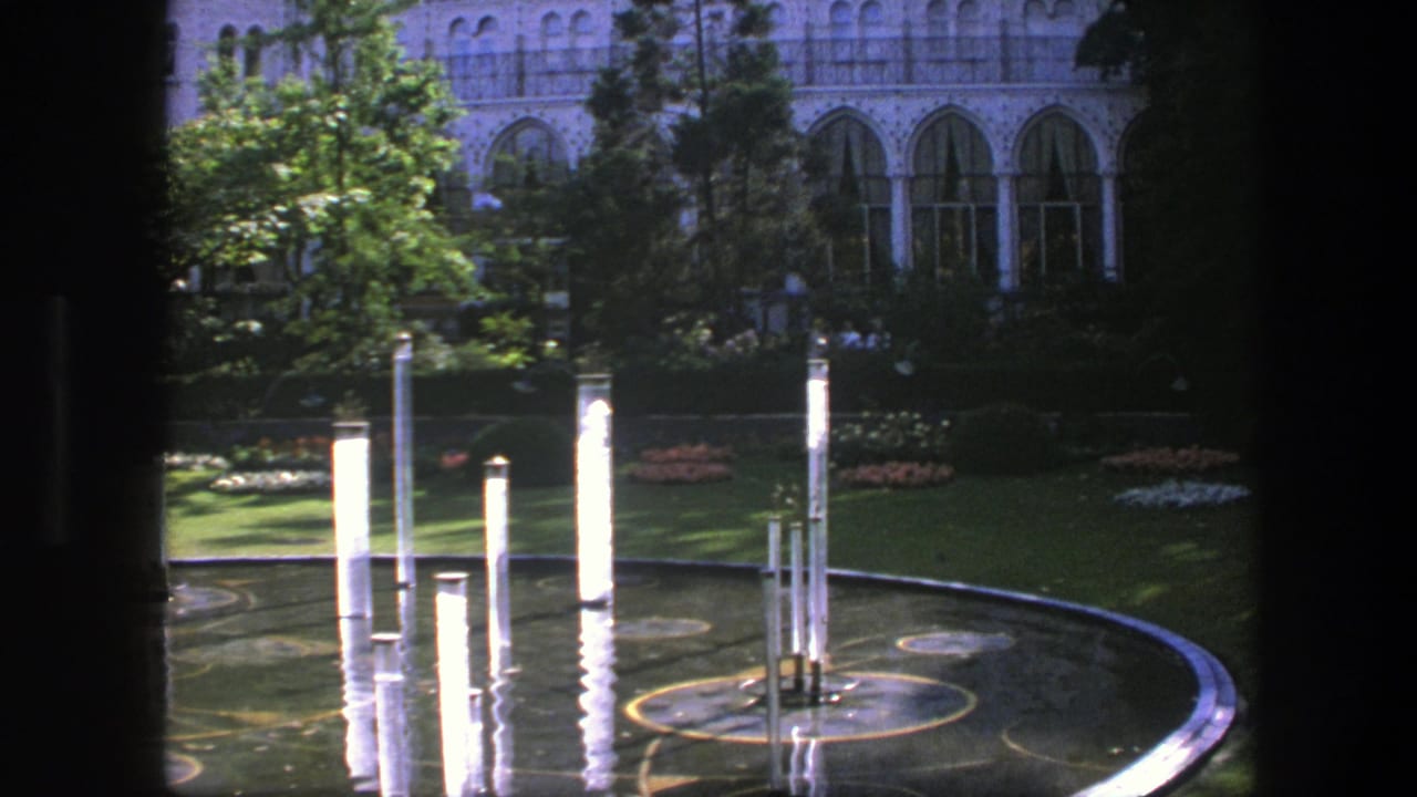 Sunlit pool edge with tall white columns, tree shadows, lawn, and arched building windows.