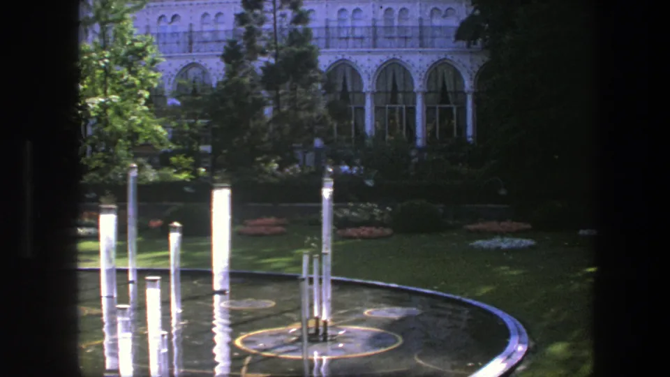 The 8mm frame holds on a Copenhagen reflecting pool, showing white columns, rippled water, lawn, shrubs, and a pale arched building.