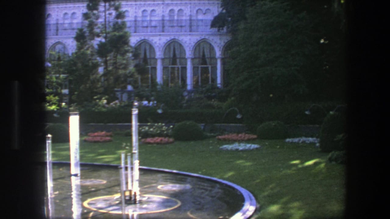 1975 Copenhagen garden view with a circular fountain, vertical water jets, lawn, flowerbeds, and an arched building facade partly shaded by trees.