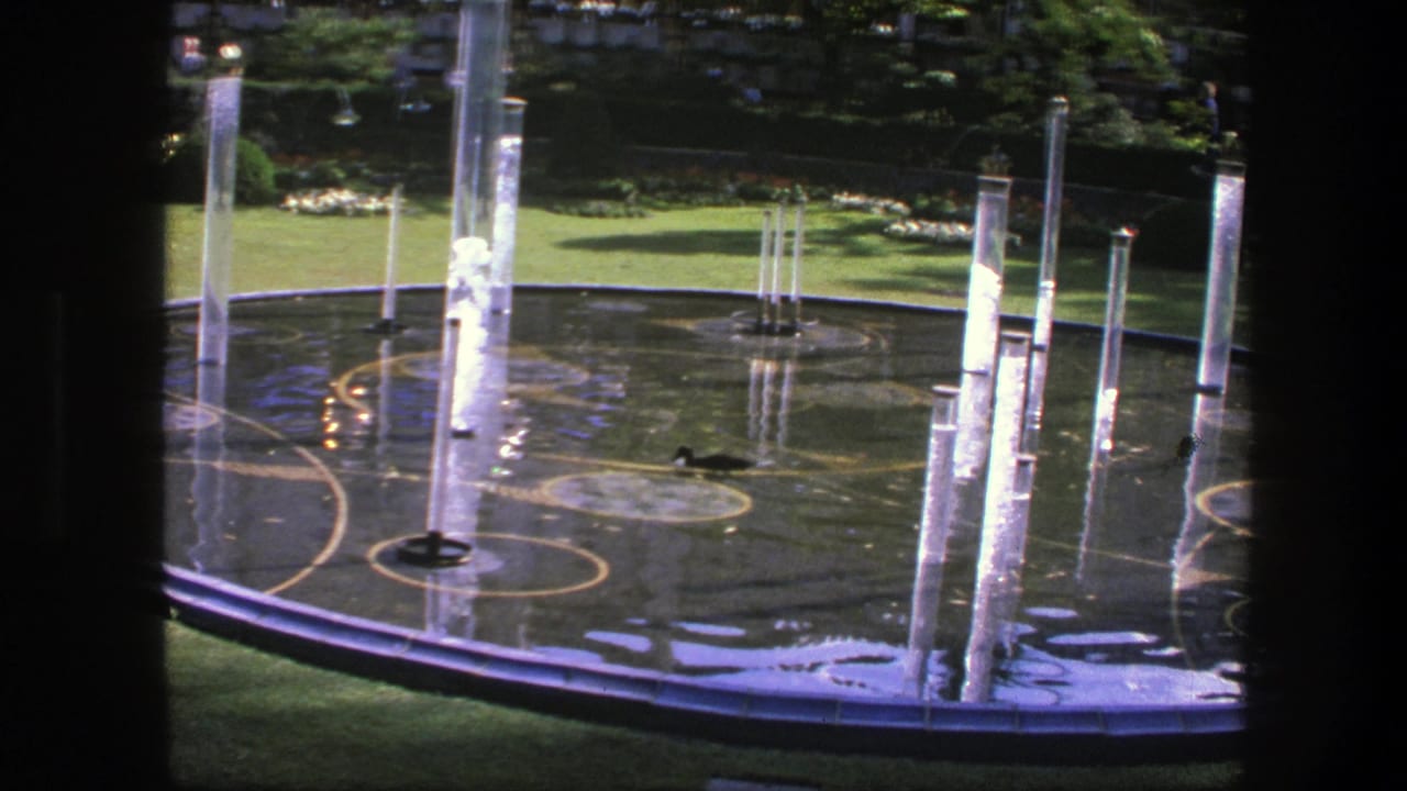 Circular garden fountain in Copenhagen, 1975, with tall clear water jets, wet reflective surfaces, surrounding lawn, and shaded planting in the background.