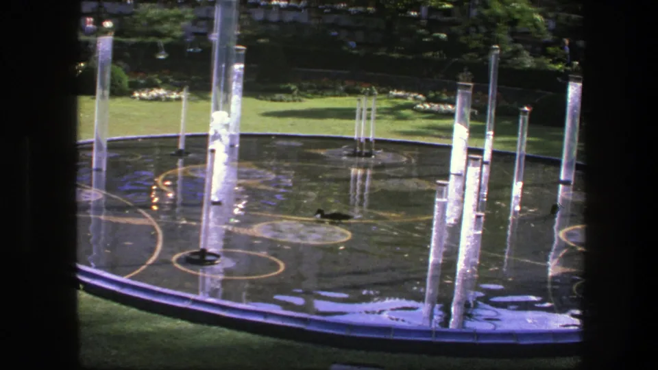 Circular garden fountain in Copenhagen, 1975, with tall clear water jets, wet reflective surfaces, surrounding lawn, and shaded planting in the background.