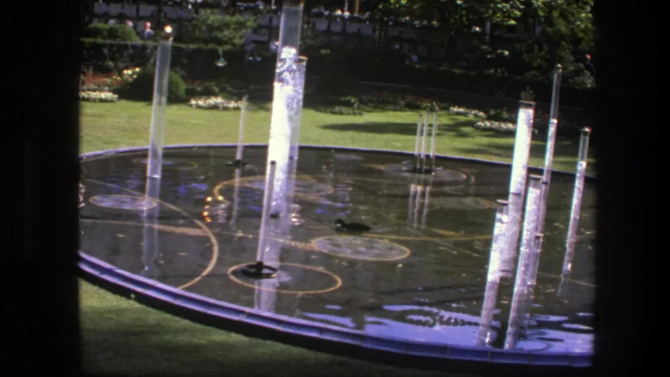 Close 1975 Copenhagen garden view of a circular fountain with tall water jets, rippled reflective surface, lawn beyond, and a small duck near the basin.