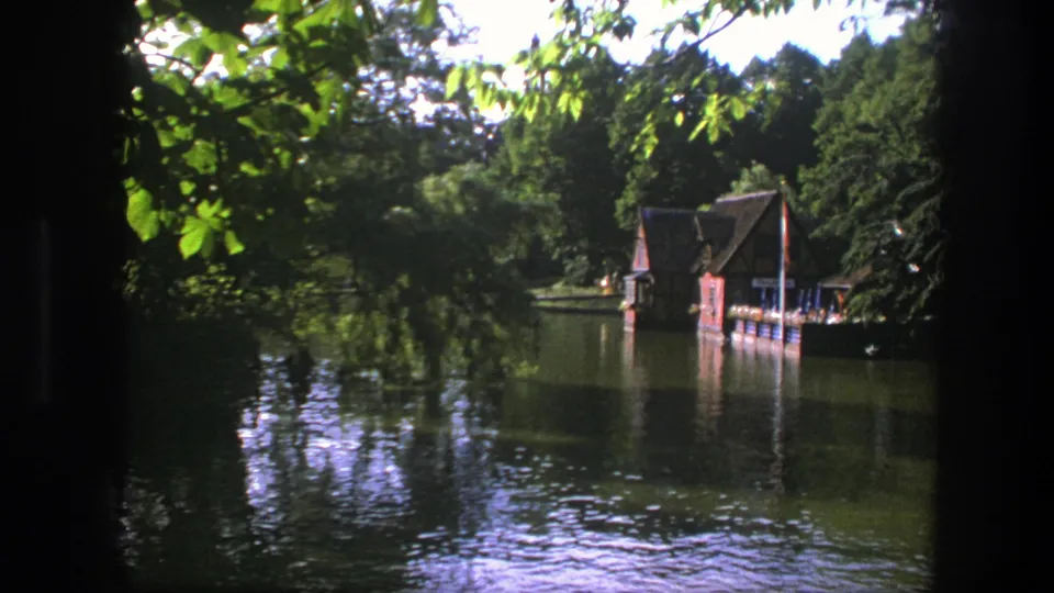 1975 Copenhagen pond scene with leafy foreground branches, dark reflective water, dense trees, and a small wooden boathouse along the shore.