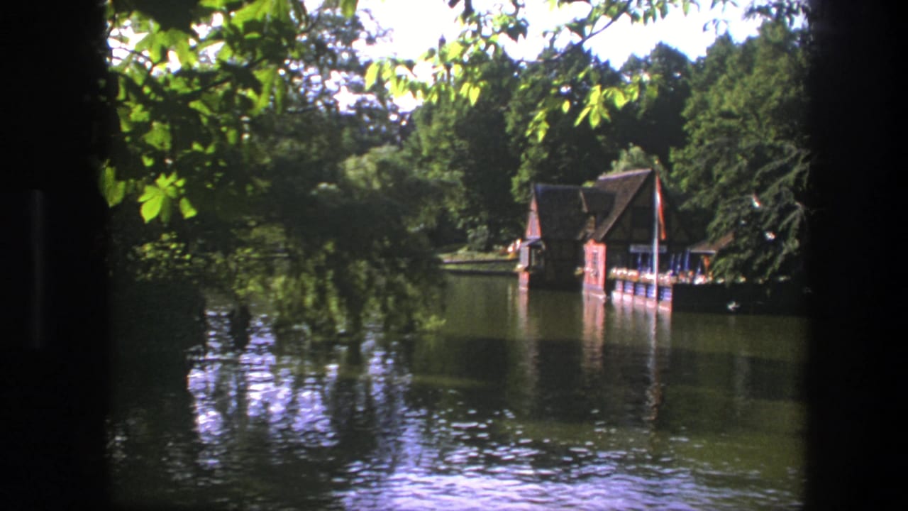 Wooden boathouse beside calm water framed by leaves and dark film edges.