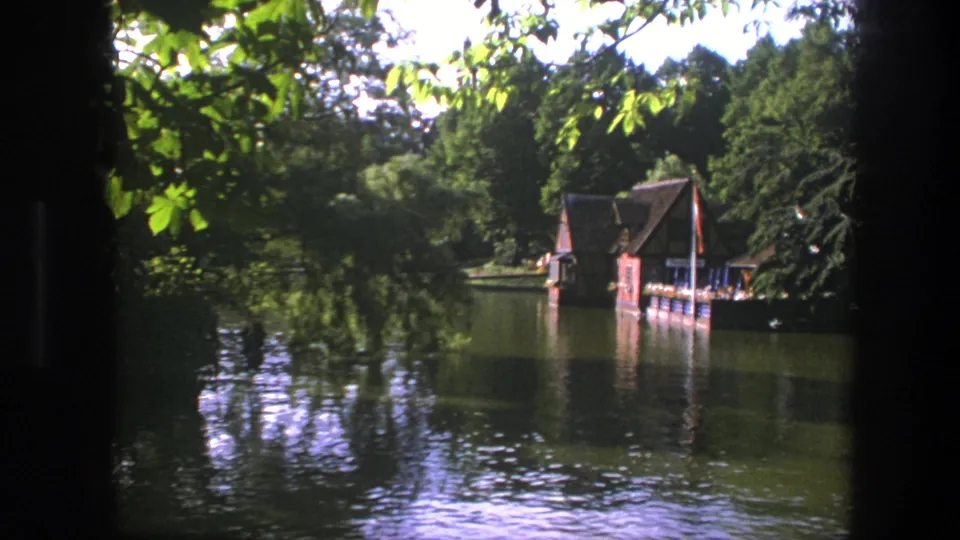 1975 Copenhagen 8mm frame showing a lakeside wooden boathouse, green trees, and reflections on calm water.