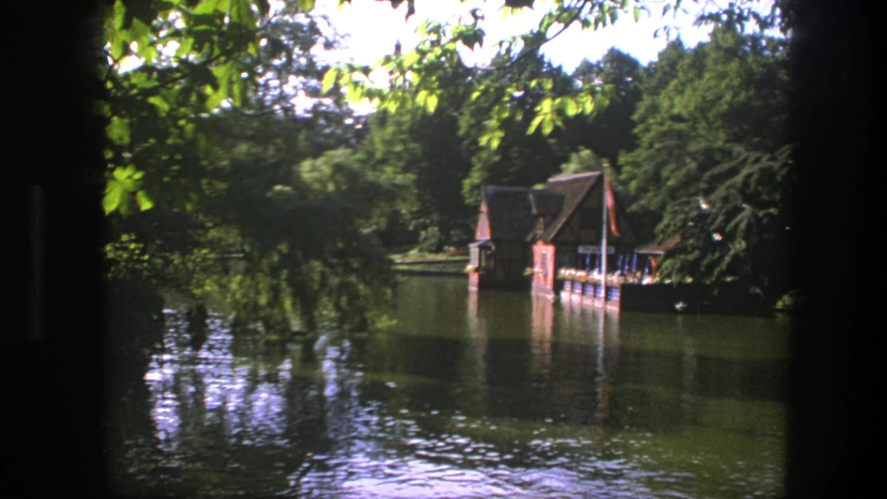 Lakeside boathouse and trees reflected in still water under bright daylight.