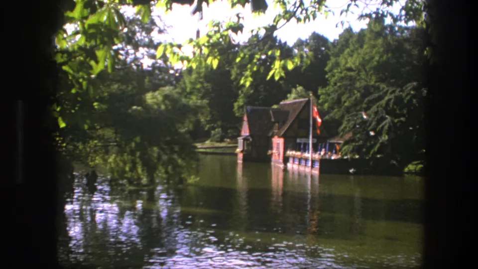 1975 Copenhagen 8mm frame with a lakeside boathouse, surrounding trees, water reflections, and a red flag near the right side.