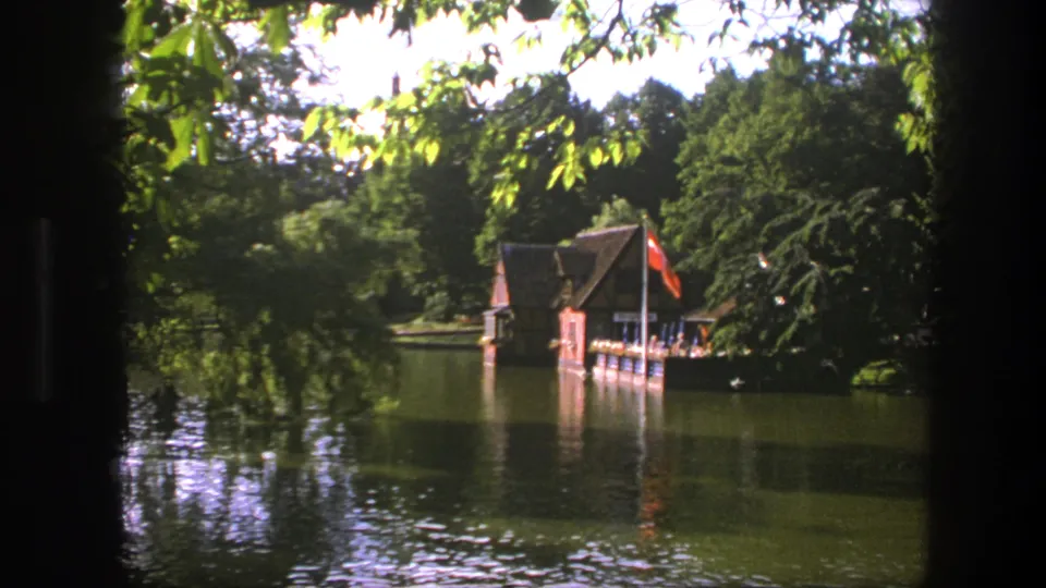 1975 Copenhagen lakeside view with a red flag, wooden boathouse, leafy branches, and calm reflective water.