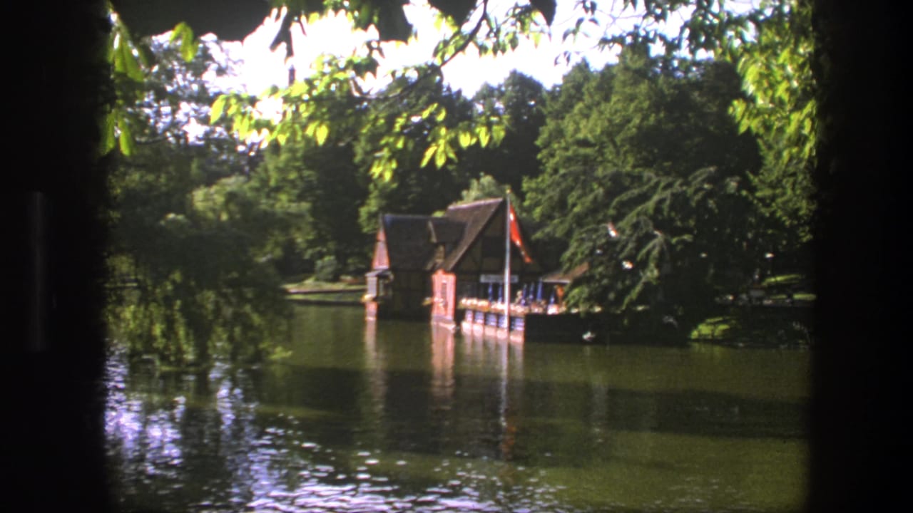 Copenhagen lakeside view on 8mm film with a boathouse, red flag, dock, trees, and rippled reflections.
