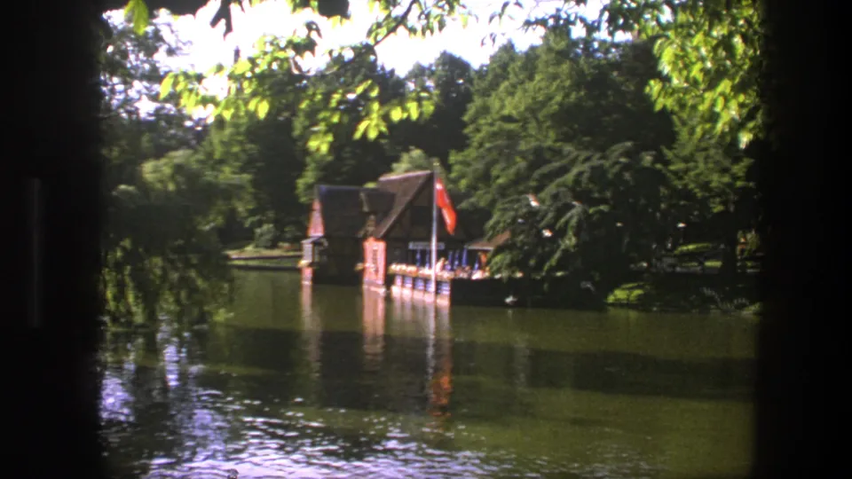 1975 Denmark home-movie frame showing a boathouse, dock, red flag, wooded shore, and reflective lake surface.