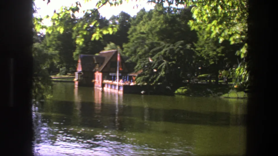 Copenhagen 1975 8mm still of a lakeside boathouse partly framed by leaves, with dock, trees, and calm water.