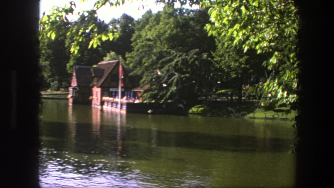 1975 Copenhagen home-movie frame of a lakeside boathouse and dock across calm water, partly obscured by dark foreground edges.