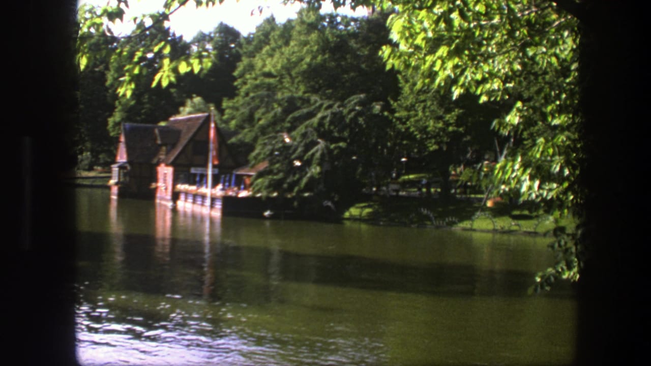 1975 Copenhagen 8mm frame showing the boathouse, dock, flag, and shaded trees reflected in a calm lake.