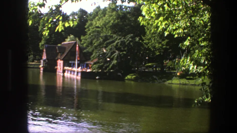 1975 Copenhagen 8mm frame showing the boathouse, dock, flag, and shaded trees reflected in a calm lake.