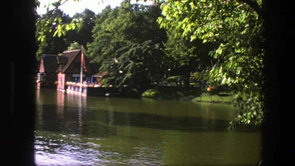 1975 Copenhagen lakeside view with a boathouse, dock, green lawn, trees, and rippled water in 8mm home-movie color.