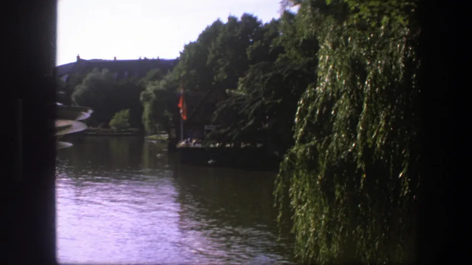 1975 Copenhagen home-movie frame showing a lagoon, dense trees, a weeping willow, and distant buildings across the water.