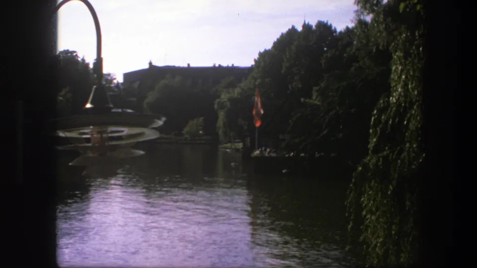 1975 Copenhagen home-movie frame showing lagoon water, a curved lamp at left, trees, and a small flagpole near the shore.