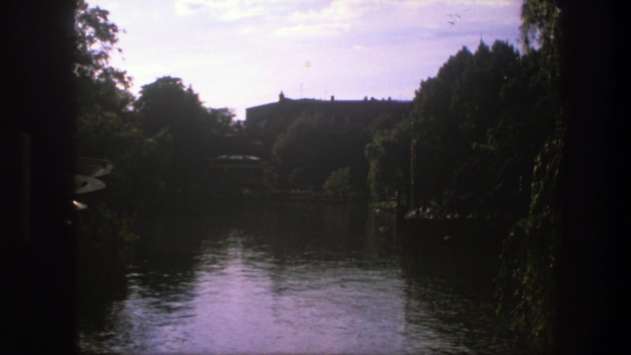1975 Copenhagen home-movie frame showing calm water, shaded trees, and a distant building across the shoreline.