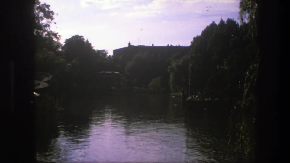 1975 Copenhagen home-movie frame showing calm water, shaded trees, and a distant building across the shoreline.