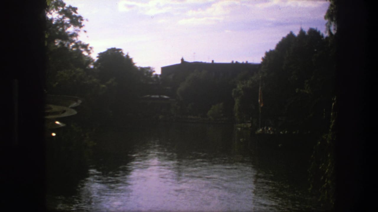 1975 Copenhagen frame with still water, dense trees, shaded edges, and a low building in the distance.