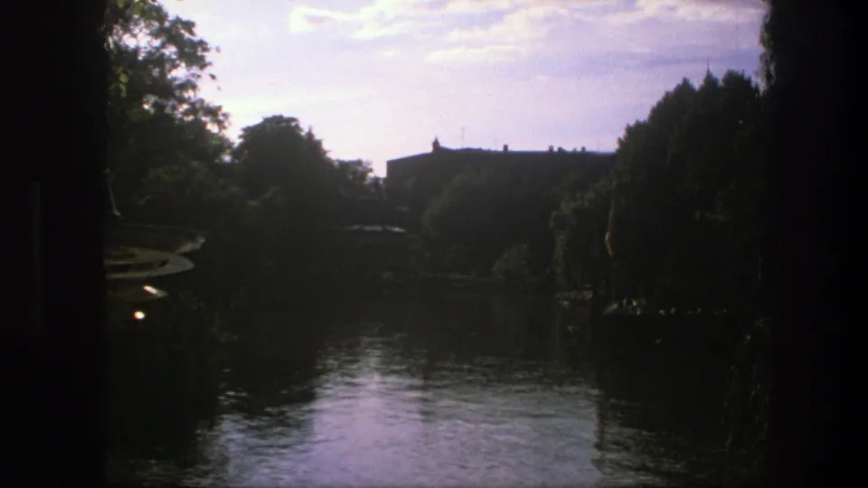 1975 Copenhagen 8mm frame showing calm water, dark trees, and a distant structure beneath a bright sky.