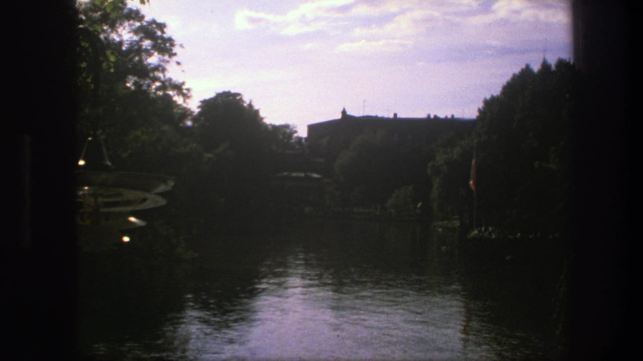 1975 Copenhagen 8mm frame showing a dark foreground view across reflective water toward trees and a distant low building.