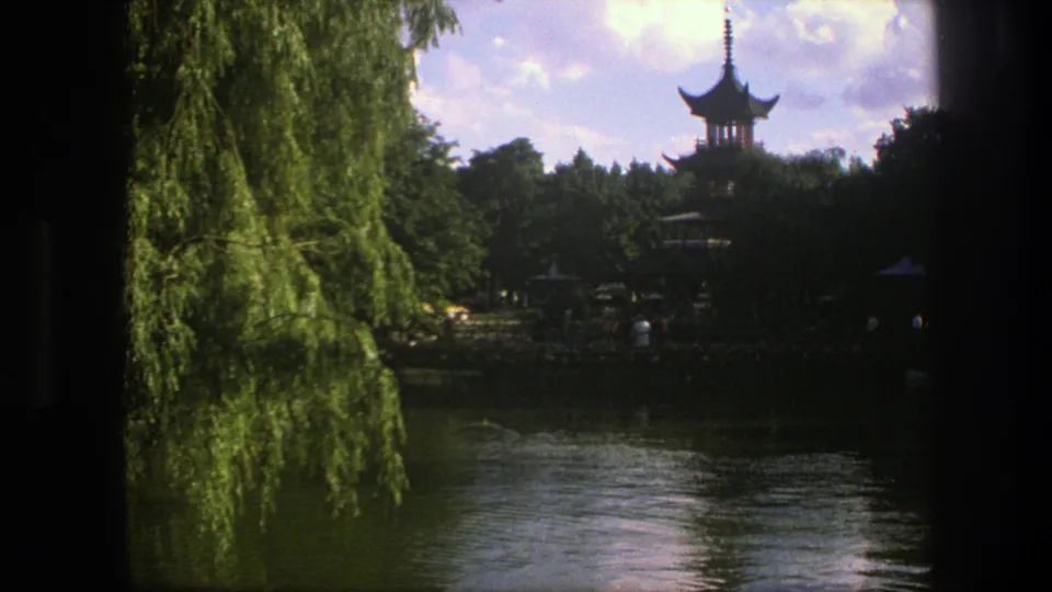 1975 Copenhagen 8mm frame showing willow branches, water, small visitors, and a distant Chinese-style pagoda above the trees.
