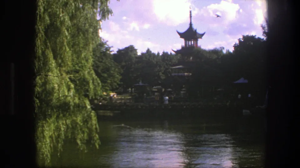 1975 Copenhagen 8mm frame with a distant Chinese-style pagoda, park trees, water reflections, tiny visitors, and a bird overhead.