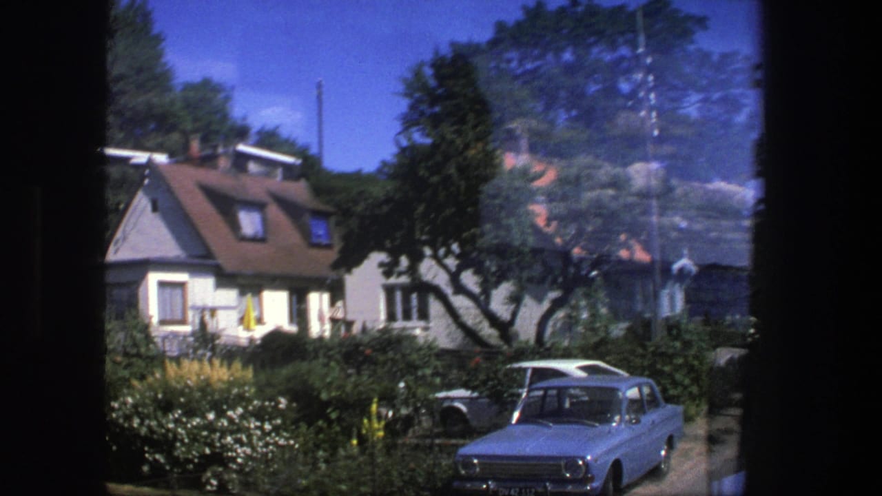 A 1975 Copenhagen neighborhood view shows a parked white car, low houses, hedges, and flowering garden beds in bright sun.