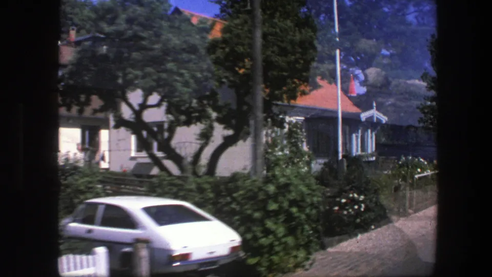 The moving 8mm camera passes a Copenhagen residential lane with a parked car, hedges, trees, and houses partly obscured by foliage.