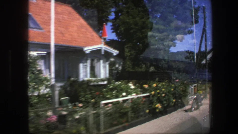 A Copenhagen house with a red roof, fenced garden, flowering plants, and roadside greenery appears in a soft 1975 8mm frame.