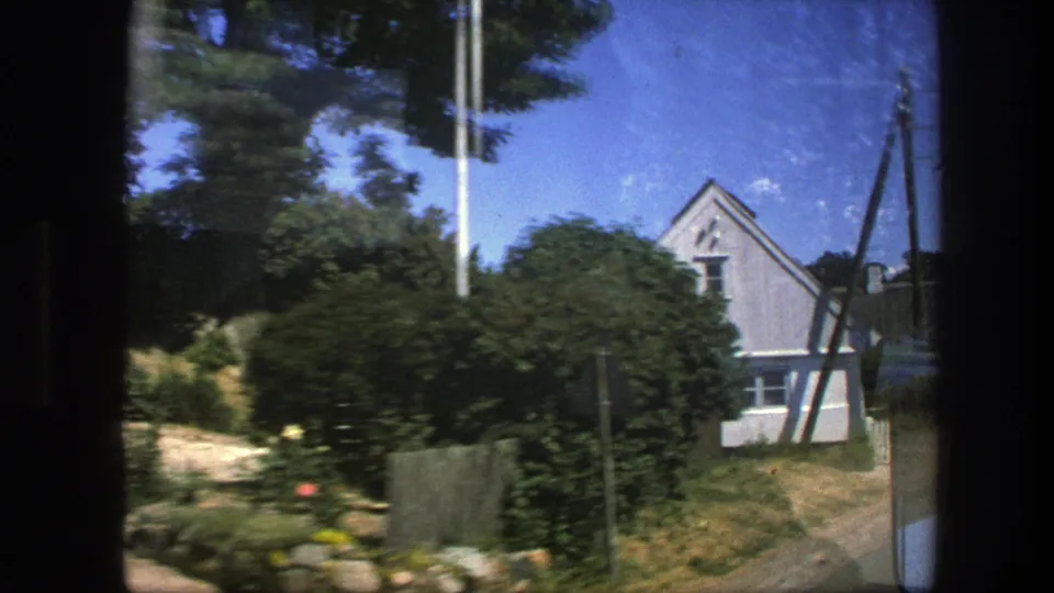 1975 Copenhagen residential view with dense garden shrubs, a pale gabled house, fencing, and roadside poles seen in motion.