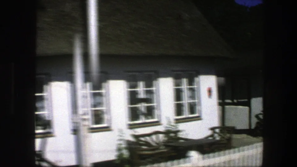 1975 Copenhagen residential frame showing a white house with a dark roof, multi-pane windows, low fence, bench, and foreground pole.