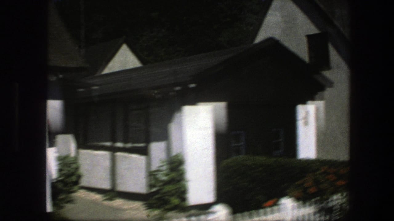 1975 Denmark home-movie frame showing a white thatched cottage with dark trim, hedges, and a small garden path.