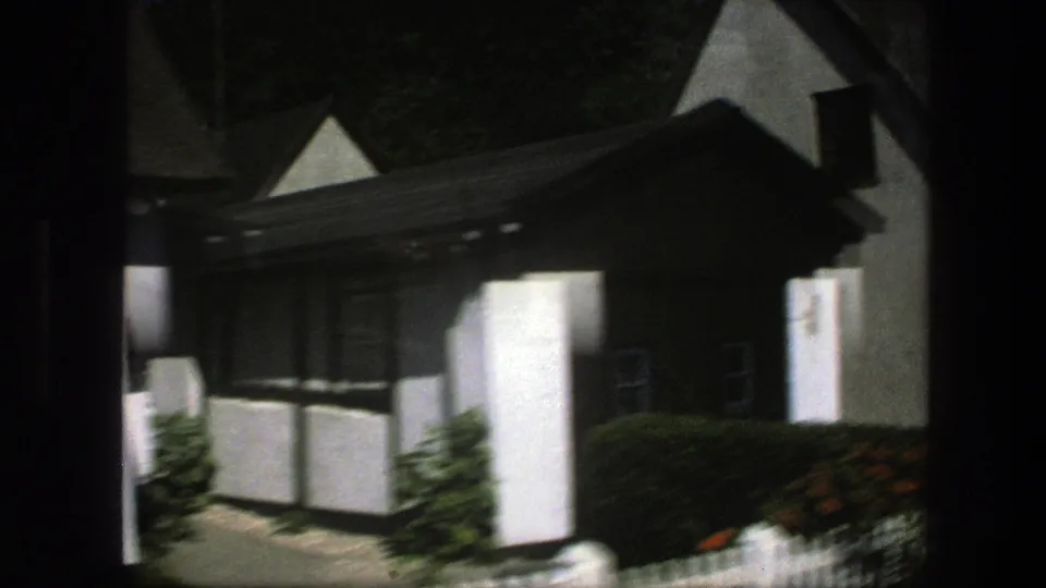 1975 Denmark home-movie frame showing a white thatched cottage with dark trim, hedges, and a small garden path.