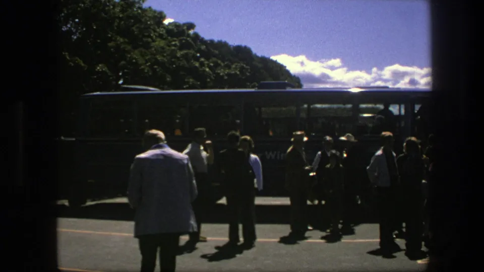 1975 Copenhagen travel frame showing tourists standing beside a coach on a sunny road with trees and sky behind them.