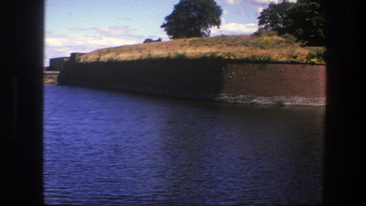 1975 Copenhagen home-movie frame showing blue moat water, a brick fortress wall, dry grass, and trees under a partly cloudy sky.