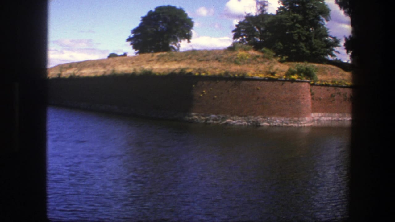 1975 Copenhagen frame with a curved brick wall along the moat, grassy bank, leafy trees, and blue sky with scattered clouds.