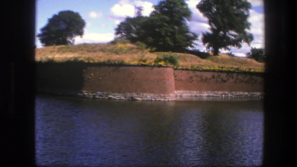 1975 Copenhagen 8mm frame showing moat water, a low brick rampart, dry grass, and large trees with dark scan borders visible.