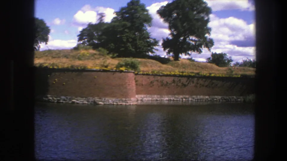 1975 Copenhagen home-movie view of a brick wall beside moat water, with trees on a grassy rise and bright clouds overhead.