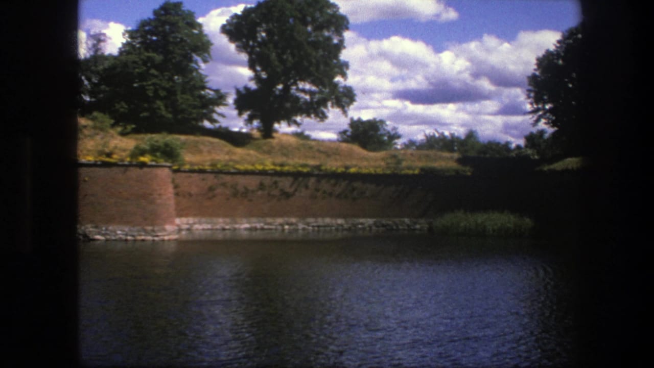 Brick wall and moat with grassy bank and trees under scattered clouds.