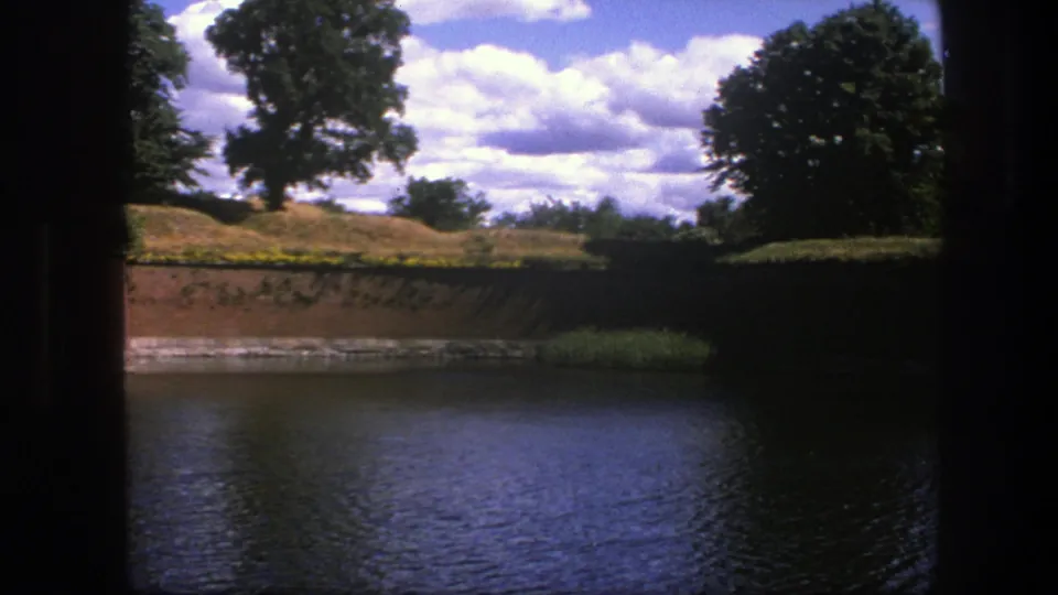 1975 Copenhagen home-movie frame with moat water in front, a brick wall and reeds along the bank, grass, trees, and clouds above.