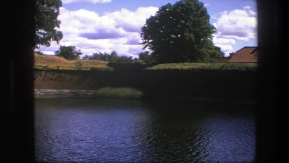 Late pan frame from Copenhagen in 1975, showing dark moat water, shaded fortification wall, reeds, trees, clouds, and a partial roofline.