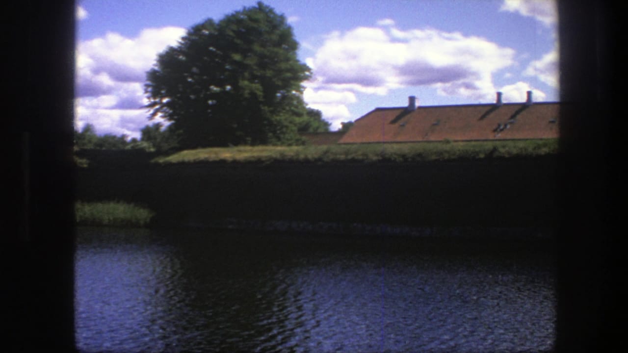 A 1975 Copenhagen waterside scene framed by dark vertical edges, showing water, a heavy wall, green tree and orange roofline.