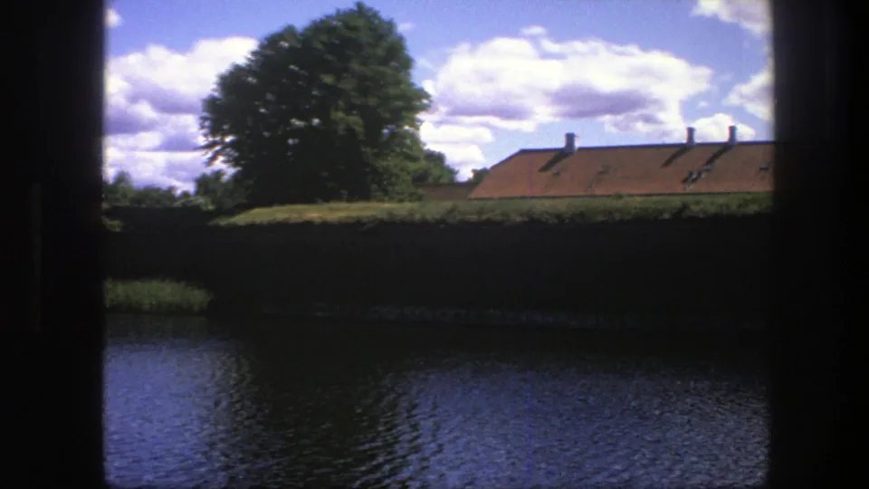 A 1975 Copenhagen waterside scene framed by dark vertical edges, showing water, a heavy wall, green tree and orange roofline.